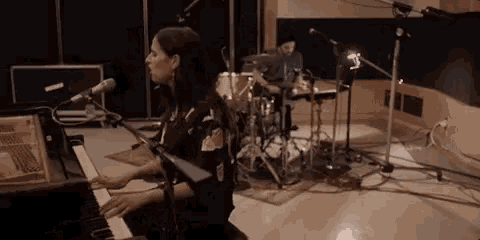 a woman is playing a piano with a man playing drums behind her