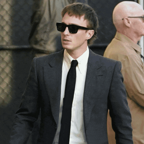 a man in a suit and tie wearing sunglasses stands in front of a chain link fence