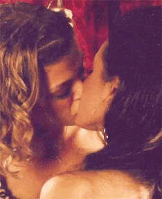 two women are kissing in front of a red curtain .