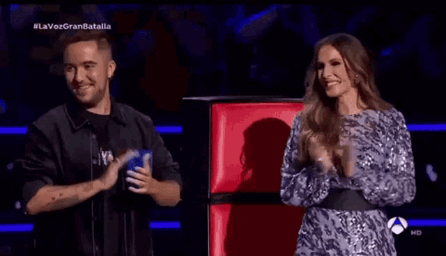 a man and a woman are clapping in front of a red chair with the words la voz gran batalla on the bottom