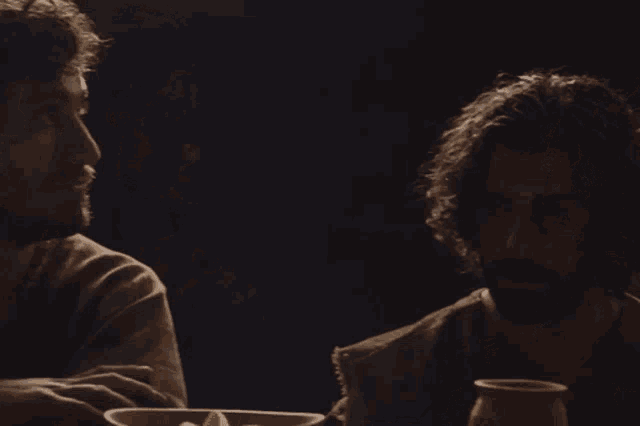 two men are sitting at a table with a bowl of food and a pitcher of wine