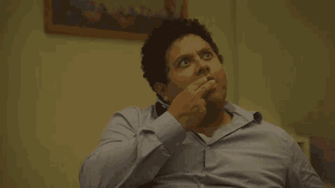 a man in a blue shirt is sitting on a couch with his hand to his chin .
