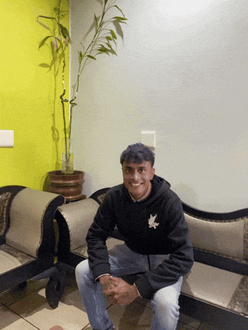 a man in a black hoodie sits on a couch next to a potted plant