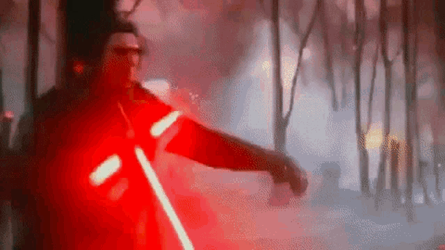 a man in a red jacket is holding a red light saber in a dark forest .