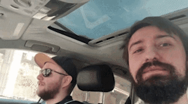 a man with a beard is sitting in the driver 's seat of a car with another man