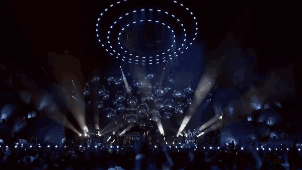 a crowd at a concert with their hands in the air and a circle of lights above them