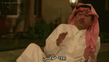 a man wearing a head scarf and a white shirt is sitting down and talking in arabic .
