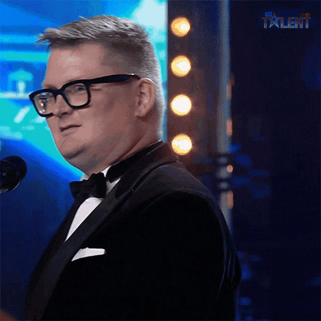 a man in a tuxedo and glasses stands in front of a microphone with the word talent on the bottom right