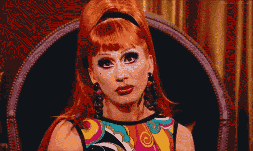 a drag queen is sitting in a chair wearing a colorful shirt and earrings .