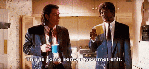 two men in suits and ties are standing in a kitchen drinking coffee and talking to each other .
