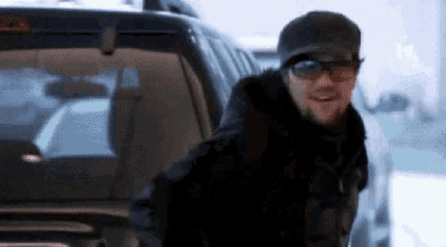 a man wearing sunglasses and a hat is standing next to a car in the snow .