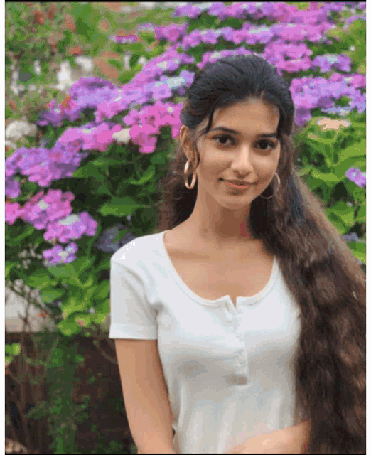 a woman in a white shirt stands in front of a bush with purple flowers
