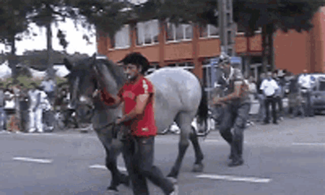 a man in a red shirt is walking a horse down a street