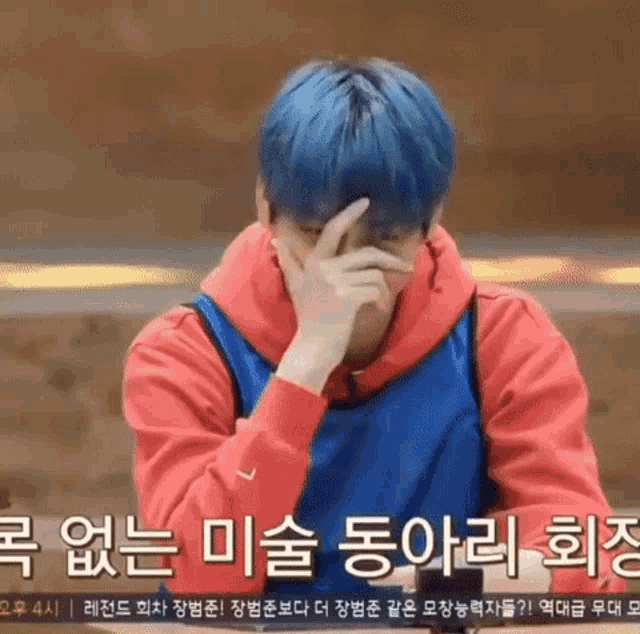 a man with blue hair and a red hoodie is covering his face with his hand