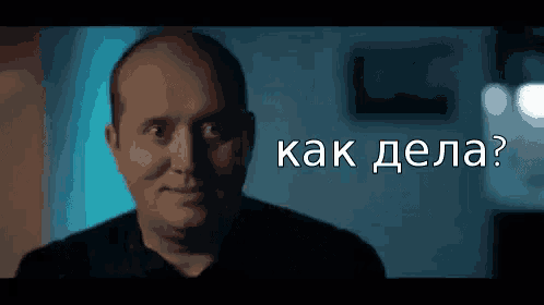 a bald man in a black shirt is looking at the camera with the words `` как дела ? '' behind him .