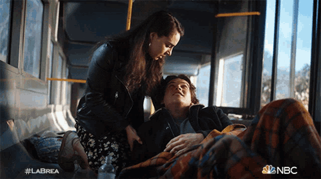 a woman kneeling down next to a man laying on a bus with the nbc logo in the corner