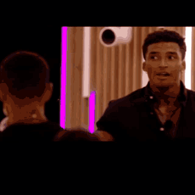 a man in a black shirt is pointing at someone in a room with purple lights behind him .