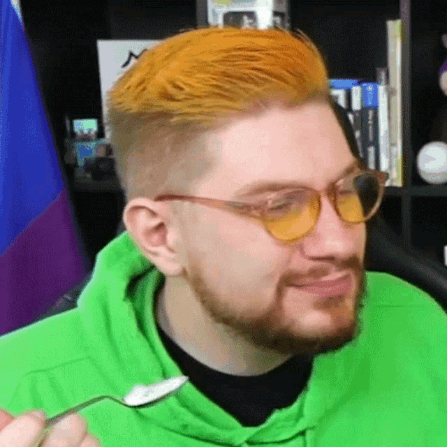 a man wearing glasses and a green hoodie is holding a spoon in his mouth .