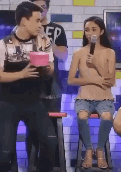 a woman sitting on a stool talking into a microphone while a man holds a box behind her