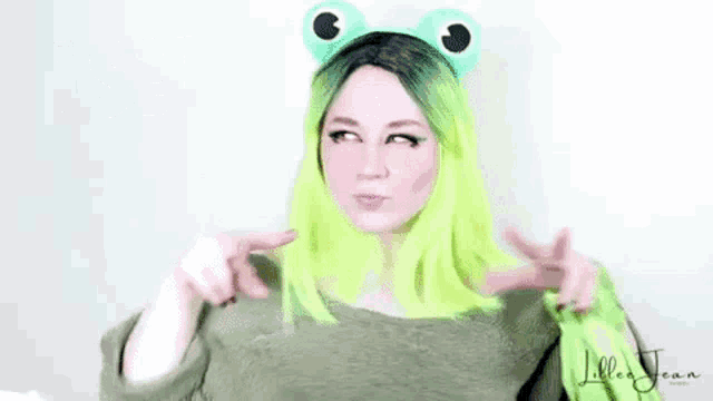 a woman with green hair is wearing a frog headband and pointing at the camera .