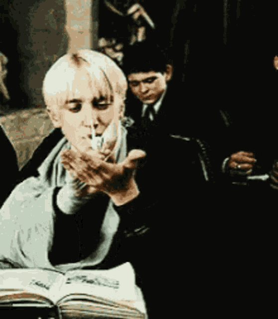 a man is smoking a cigarette while sitting at a table with a book open .