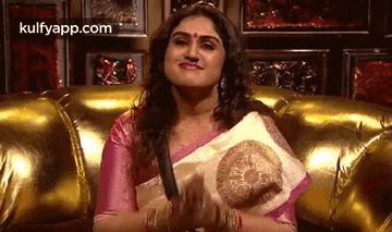 a woman in a pink saree is sitting on a gold couch holding a white pillow .