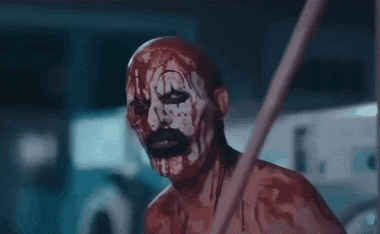 a bloody clown with blood on his face is waving his hands in a dark room .