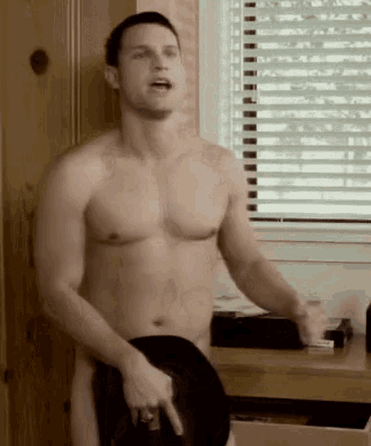 a shirtless man is standing in front of a window and holding a hat .