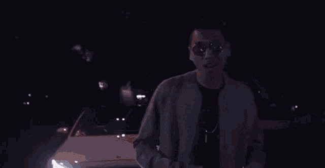 a man wearing sunglasses is standing in front of a car in the dark .