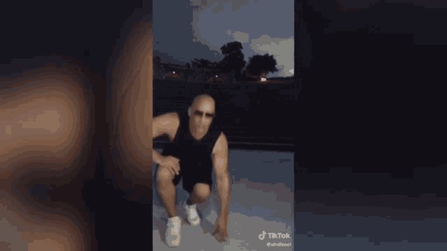 a man wearing sunglasses and a black tank top is squatting down on a concrete surface .