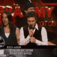 a man is speaking into a microphone in front of a sign that says rock album stadium arcadia red hot chili peppers