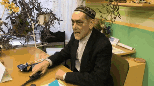 a man in a suit and hat sits at a desk holding a gun