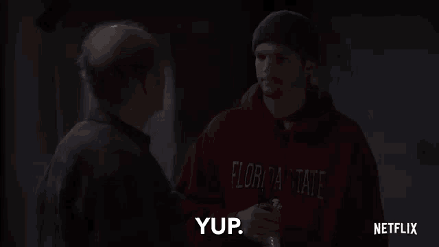 a man in a red florida state hoodie is talking to another man in a dark room .