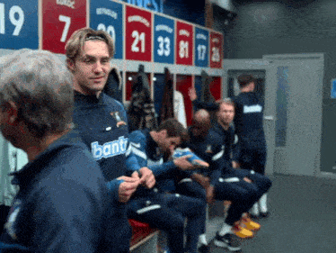 a man wearing a bant shirt sits in a locker room with other players