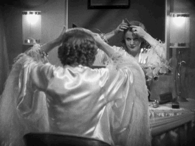 a black and white photo of a woman getting ready in front of a mirror
