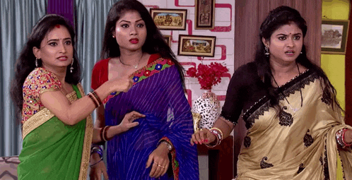 three women are standing next to each other in a room . one of the women is wearing a blue saree .