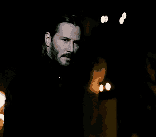 a man with long hair and a beard is standing in the dark with a serious look on his face .