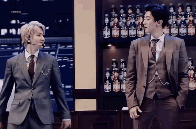 two men in suits and ties are standing next to each other in front of a shelf full of bottles .