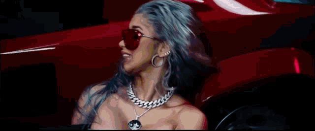 the woman is wearing sunglasses and a necklace and is standing in front of a red car .