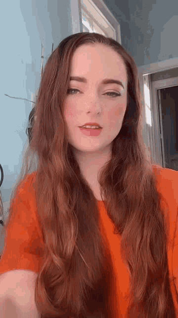 a woman with long red hair and freckles is wearing an orange shirt and standing in front of a window .
