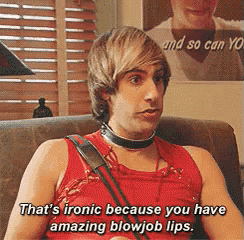 a man in a red tank top is sitting on a couch and says that 's ironical because you have amazing blowjob lips