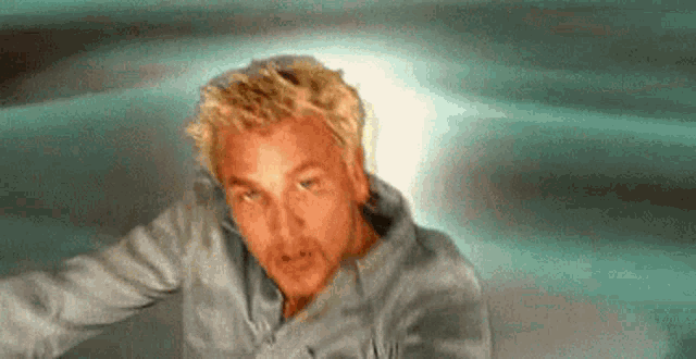 a man with blonde hair and a blue shirt is standing in front of a blue background .