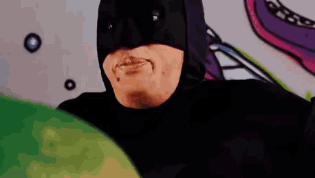a man in a batman costume is standing in front of a wall with graffiti on it .