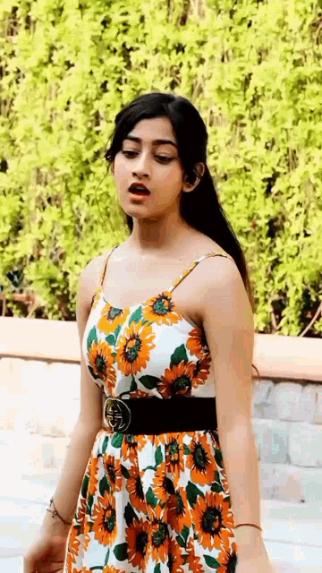 a woman in a floral dress is standing in front of a green bush .