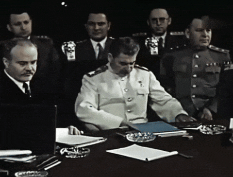 a man in a white uniform sits at a desk in front of a group of men in military uniforms