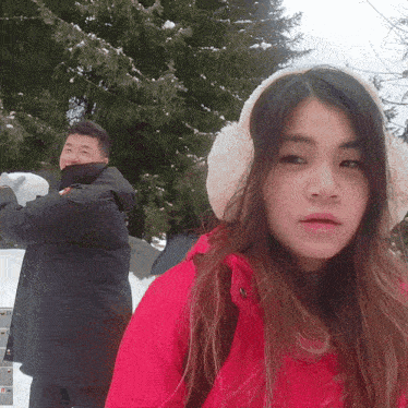 a woman wearing ear muffs looks at the camera while a man throws a snowball behind her