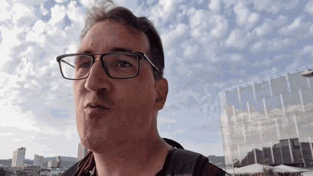 a man wearing glasses stands in front of a building with a blue sky in the background