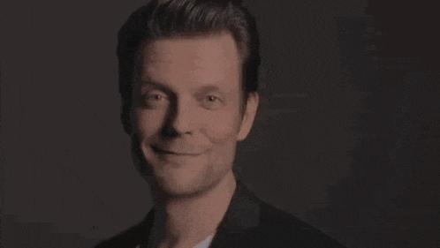 a man in a suit and white shirt is smiling and looking at the camera in a dark room .