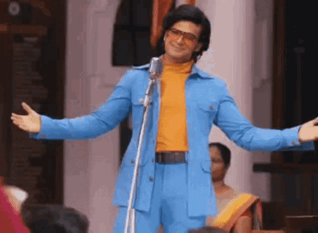 a man in a blue suit and orange turtleneck is standing in front of a microphone with his arms outstretched .