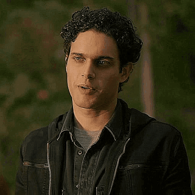 a man with curly hair wearing a black jacket and a denim shirt is looking at the camera .